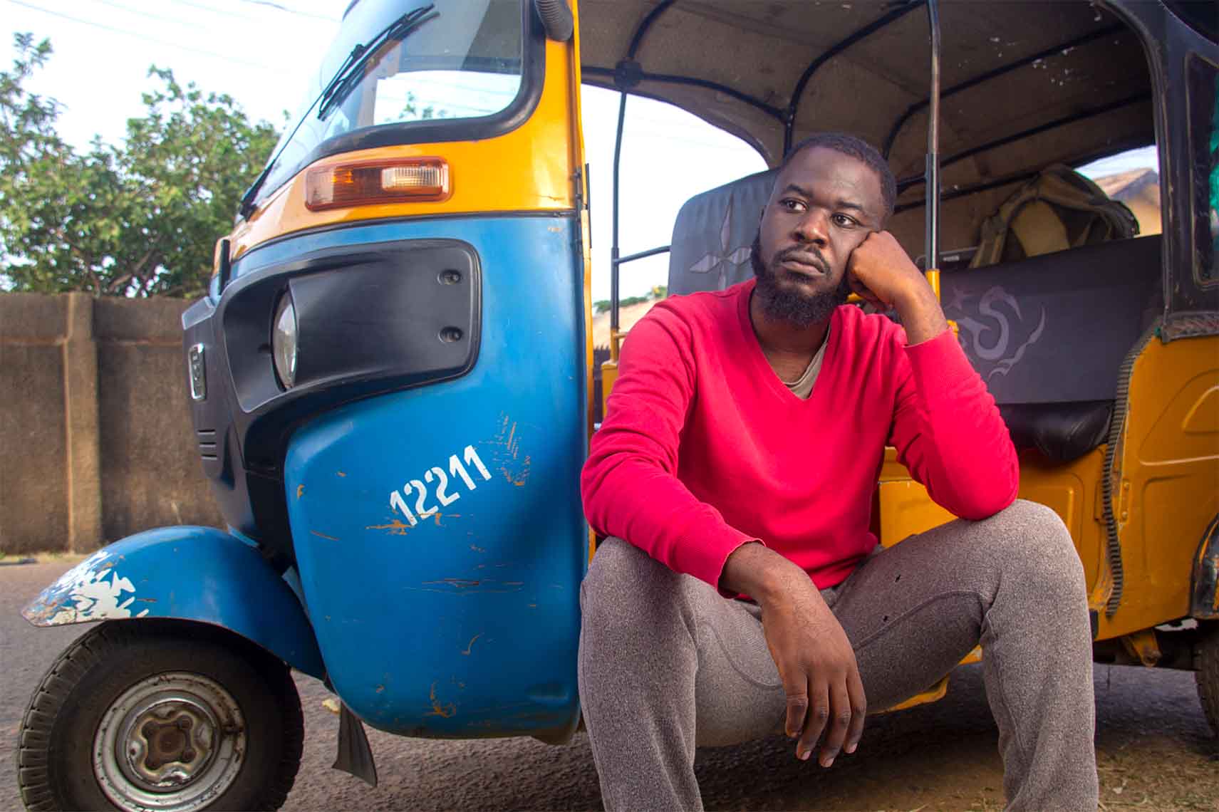 A worried African tricycle keke maruwa rider standing in front of his tricycle, with both hands on his cheeks, looking sad 2.